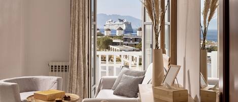 A modern hotel room with a view of a cruise ship, a balcony with pampas grass, and a cozy seating area with a table and chairs.