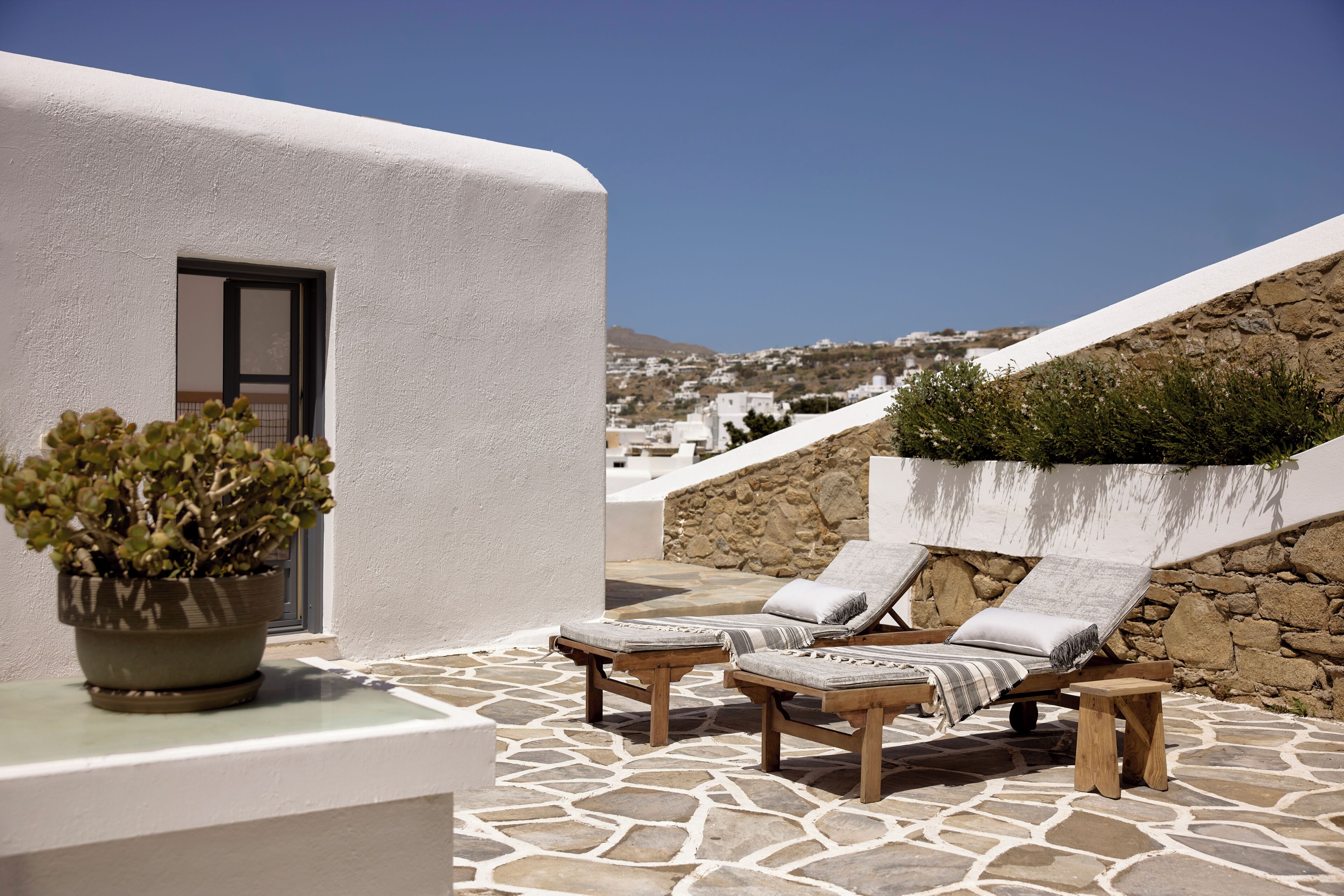 A terrace with stone flooring, two wooden lounge chairs with cushions, a potted plant, and a view of a white building and hillside in the background.