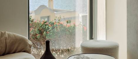 A room with a sofa, a small round table, a vase, and a view of a building and greenery through a window.