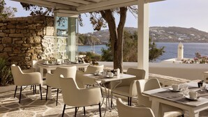 Una terraza con muros de piedra, mobiliario blanco y vistas al mar y una colina con edificios.