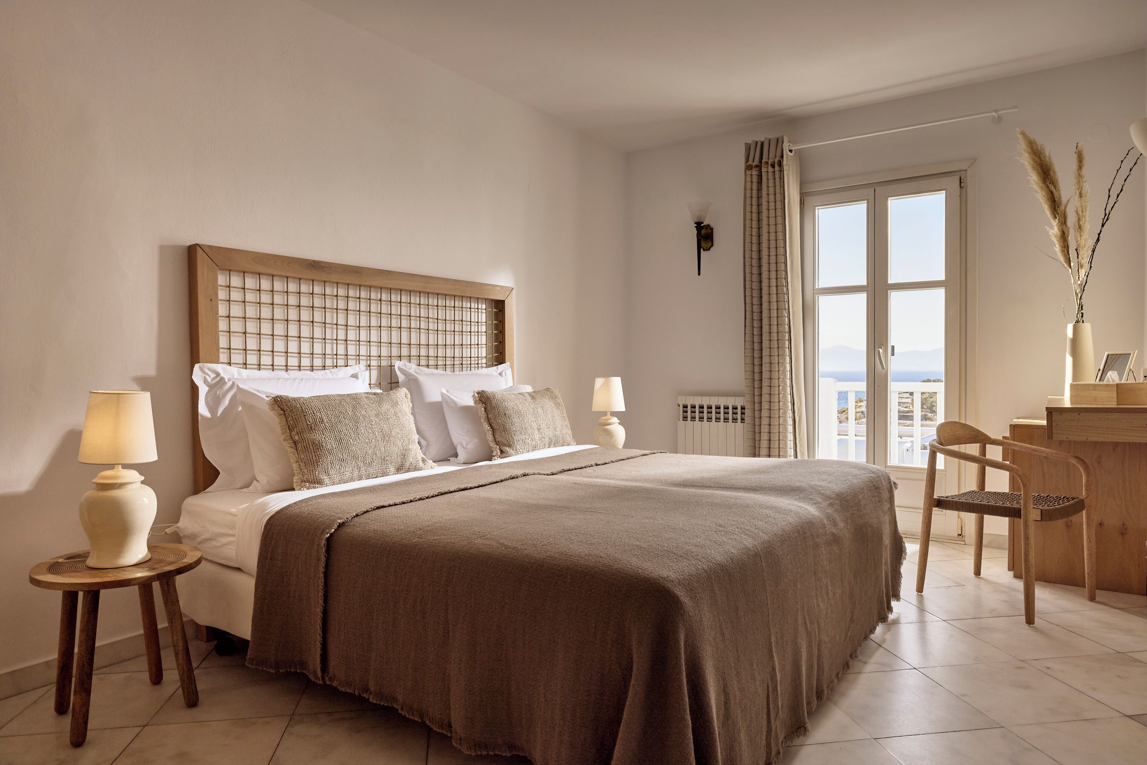 A bedroom with a large bed, two bedside tables with lamps, a wooden desk, and a view of the outdoors through a window.