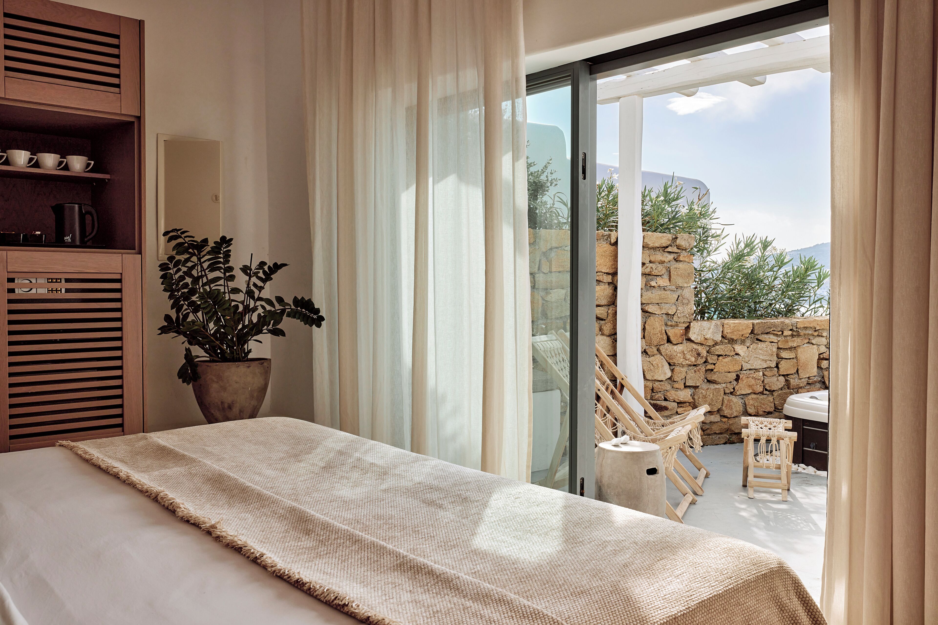 A bedroom with a bed, a potted plant, a wooden shelf, and a view of a stone wall and outdoor seating area.