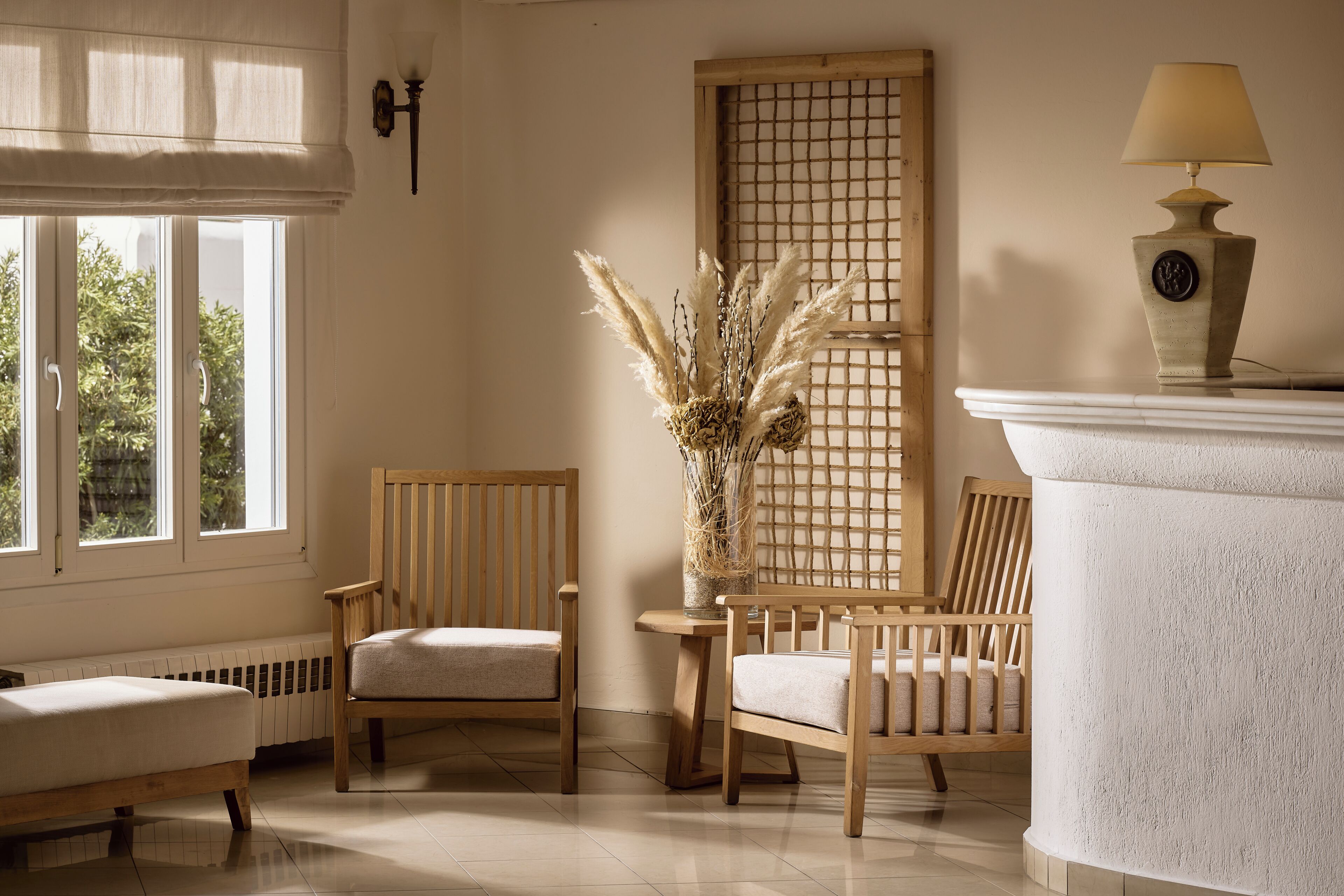 A room with two wooden chairs, a small table, a vase with pampas grass, a lamp, and a window with a view of greenery.