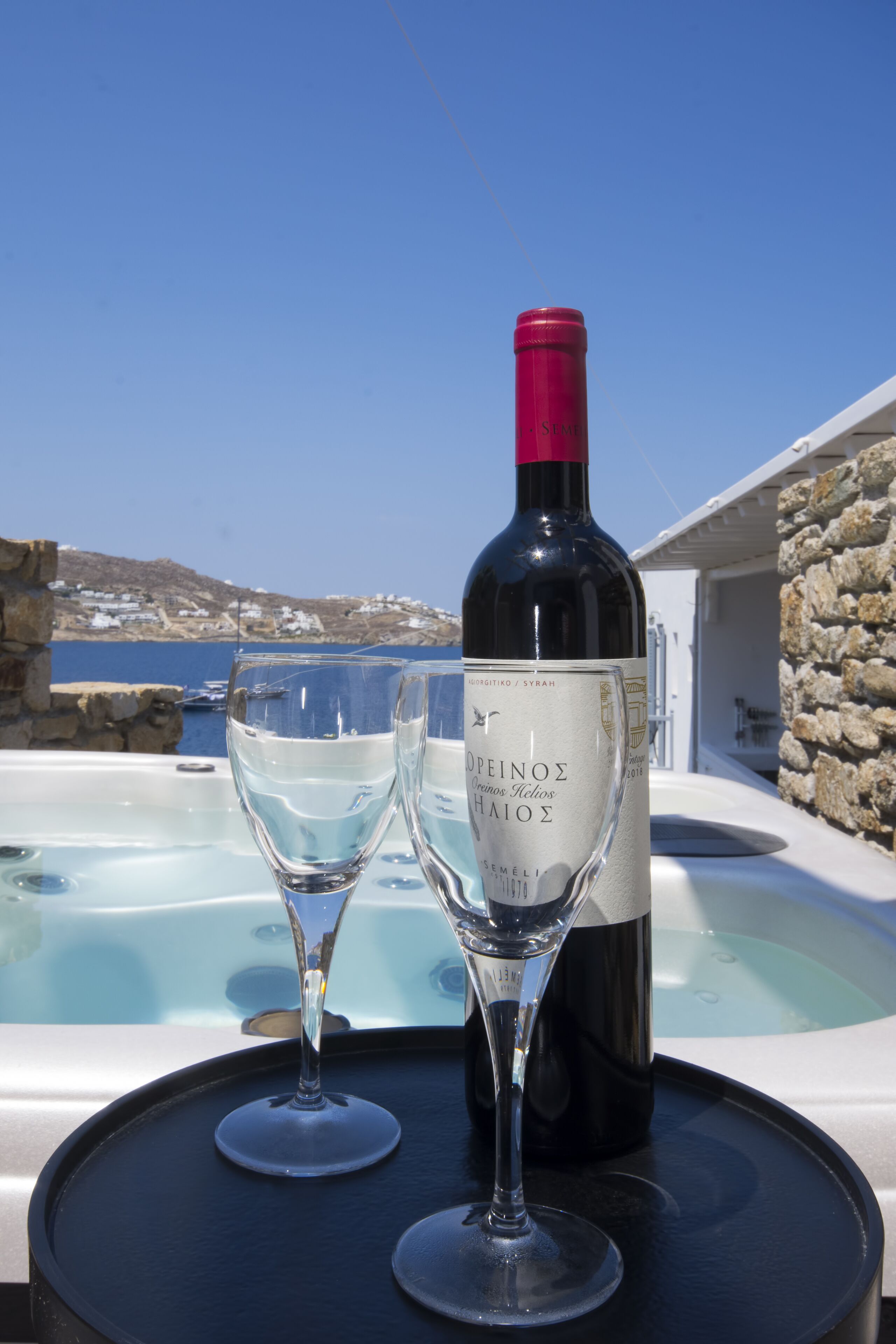 A bottle of wine and two glasses on a table with a hot tub and a scenic view of the sea and buildings in the background.