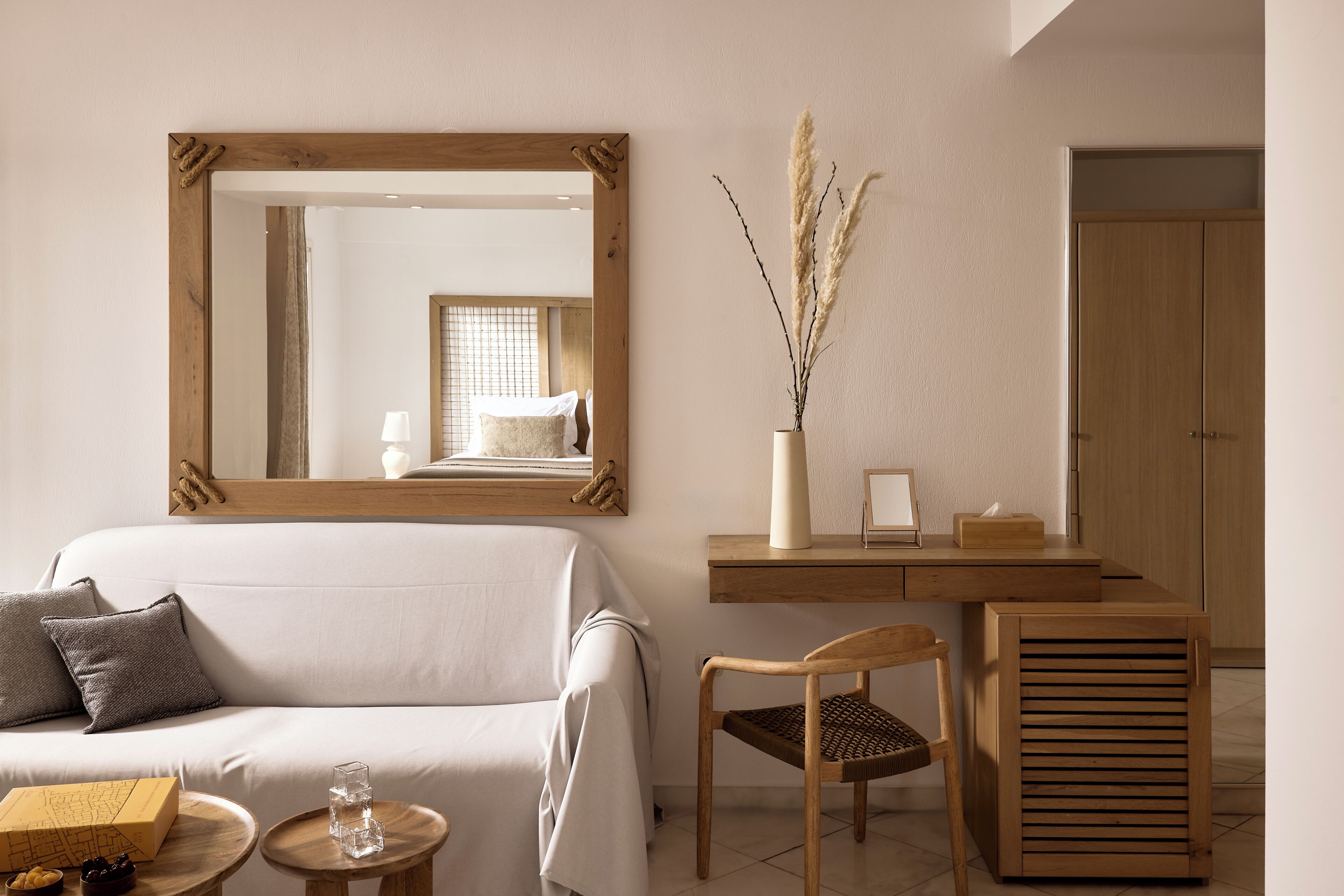 A room with a sofa, a wooden chair, a small round table with a glass and a box, a wooden desk with a vase, and a large mirror on the wall.