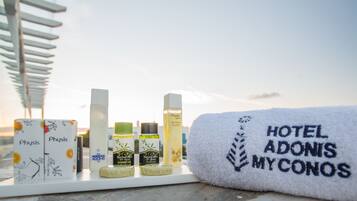 Un baño de hotel con un perchero, distintos artículos de aseo y una toalla bordada con la inscripción 'HOTEL ADONIS MYKONOS'.