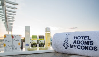 Un baño de hotel con un perchero, distintos artículos de aseo y una toalla bordada con la inscripción 'HOTEL ADONIS MYKONOS'.