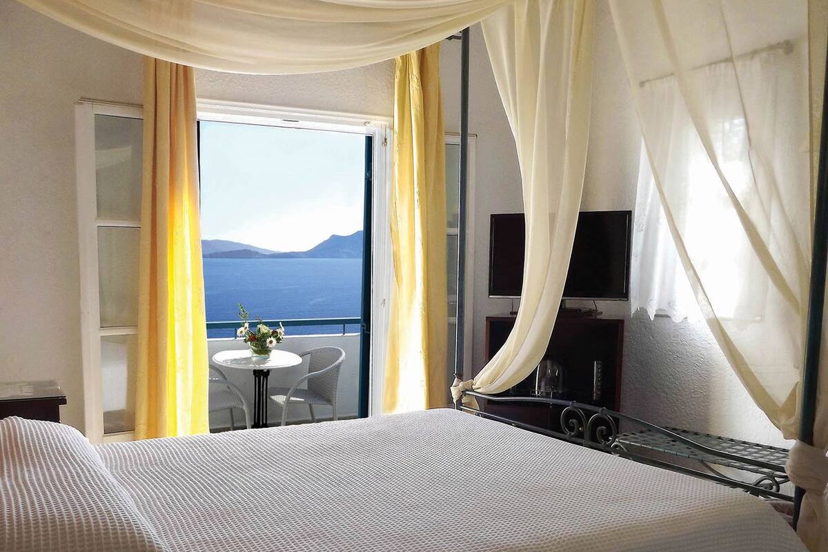 A hotel room with a bed, a TV, a balcony with a table and chairs, and a view of the sea.