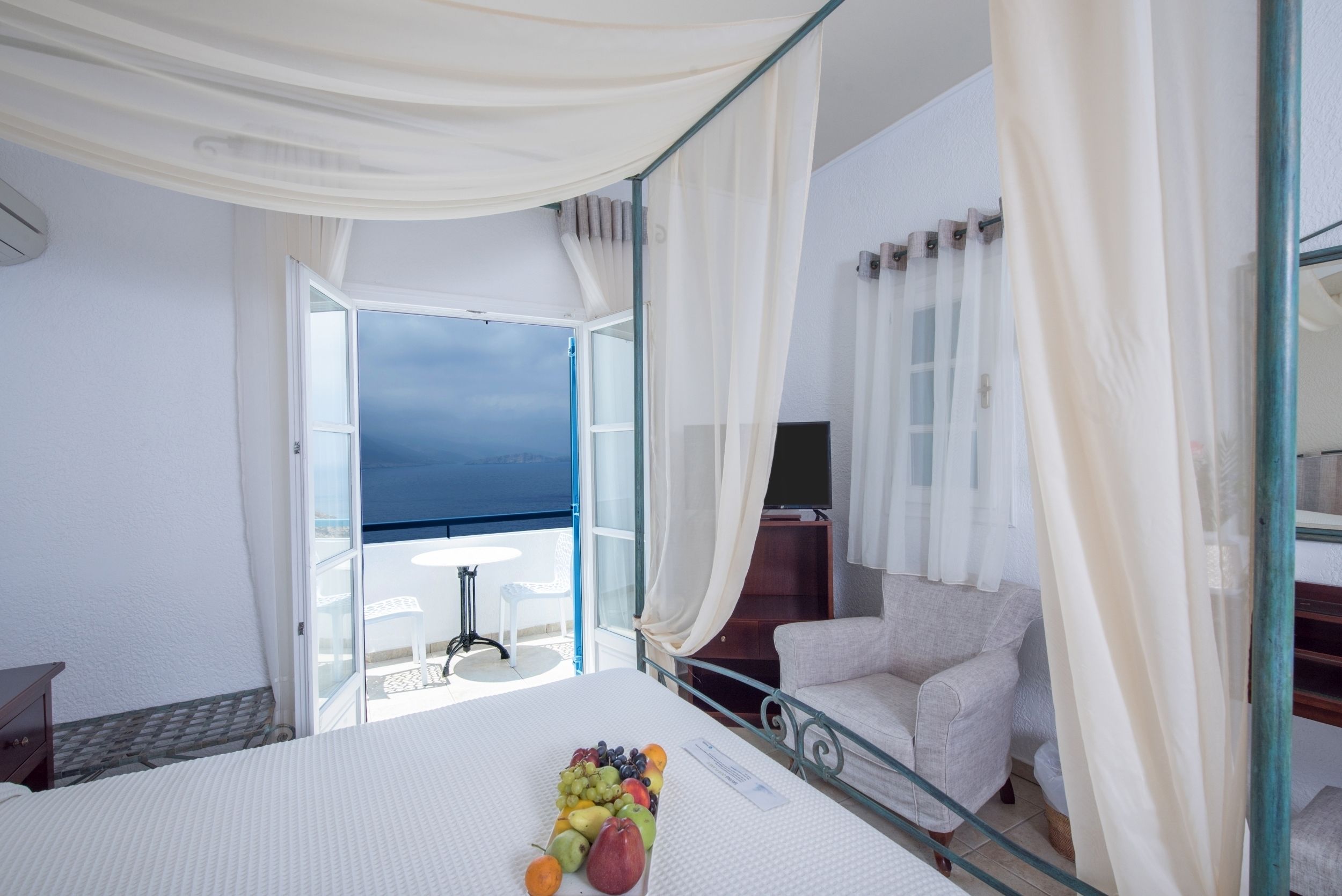 Habitación de hotel con cama, cesta de frutas, una silla y balcón con vistas.