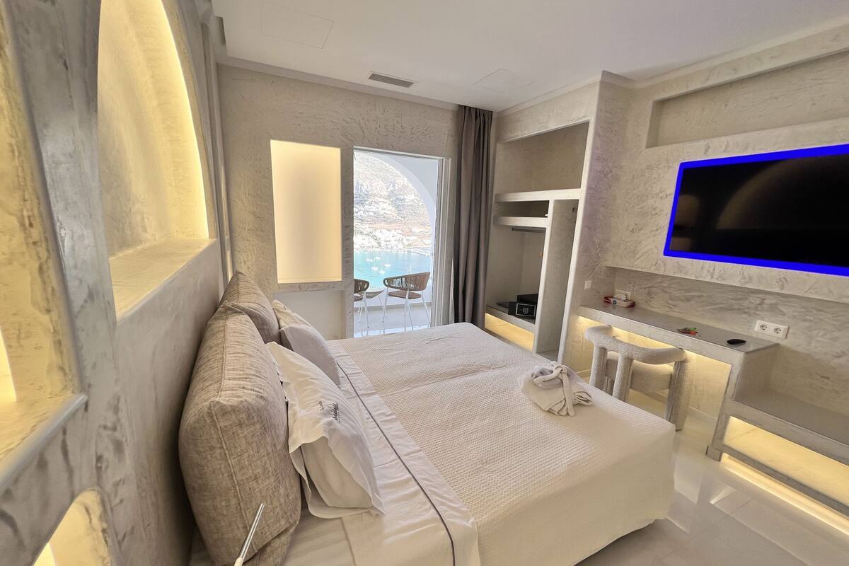 A modern hotel room with a large bed, a TV mounted on the wall, and a balcony with a view of the sea.