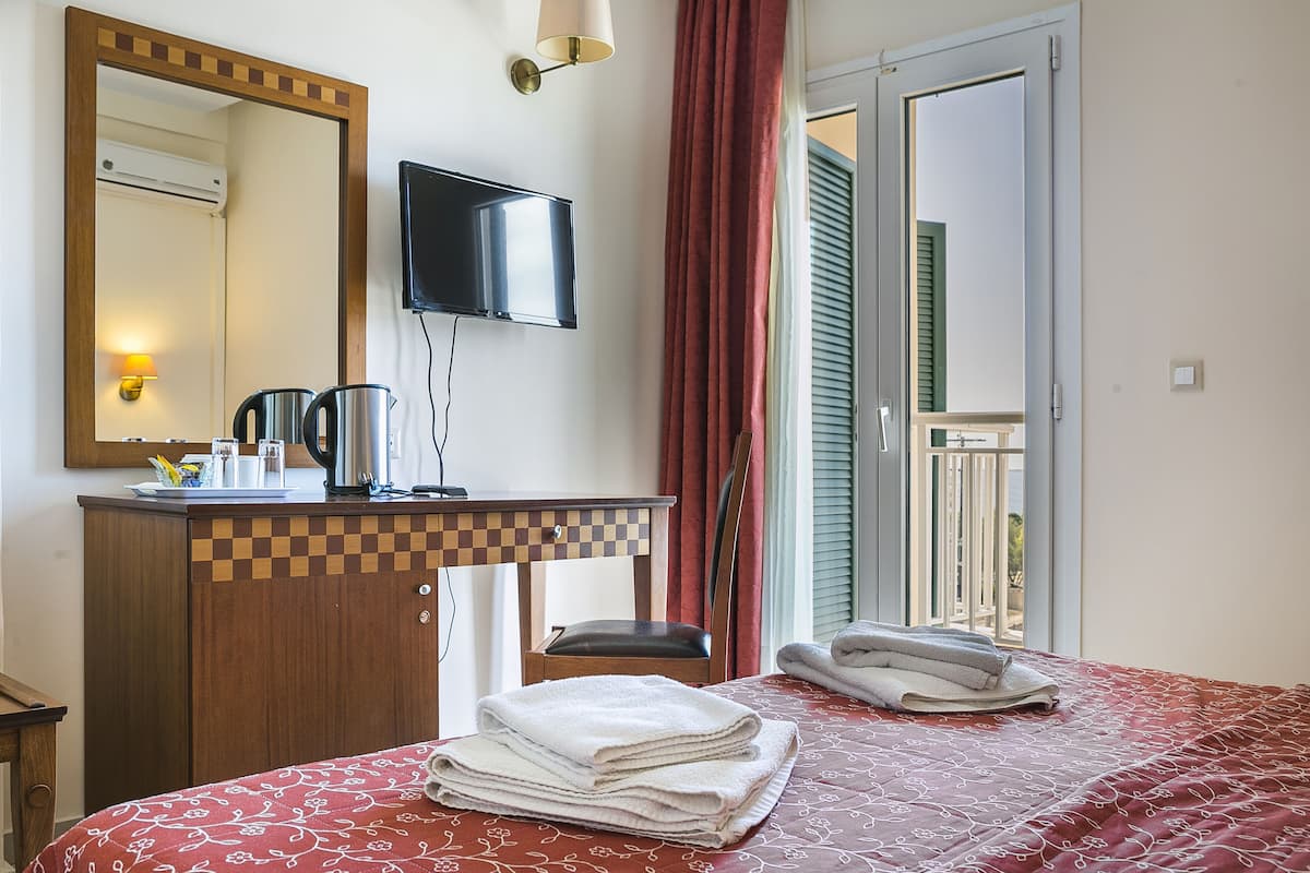 Habitación de hotel con una cama, un escritorio con televisor de pantalla plana, cafetera y una silla. Hay toallas sobre la cama y una ventana con cortinas.