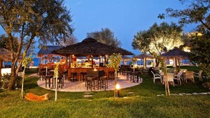 Un área de comedor al aire libre con mesas y sillas, rodeada de árboles y una estructura con techo de paja.