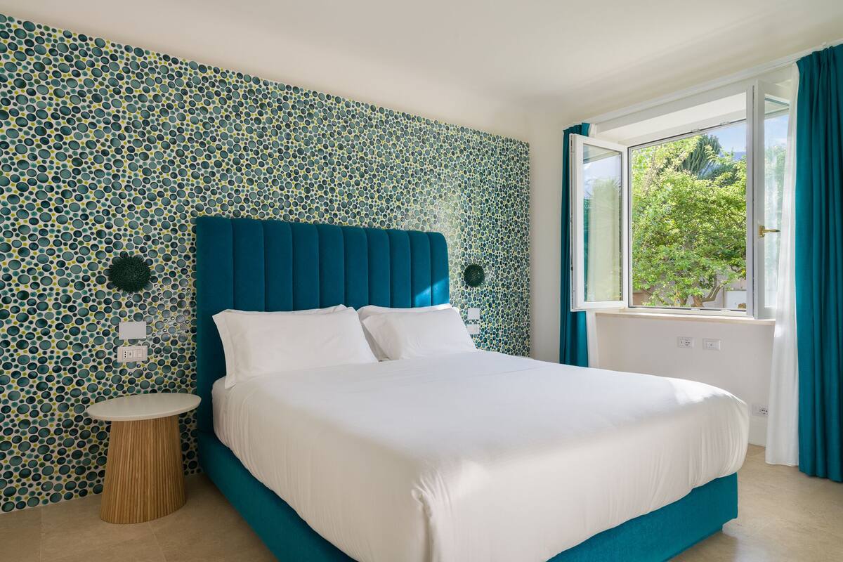 A neatly made bed with a blue headboard in a room with patterned wallpaper and a window with curtains.