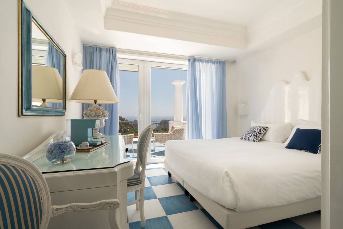 A hotel room with a bed, a desk, a chair, a lamp, and a view of the sea.