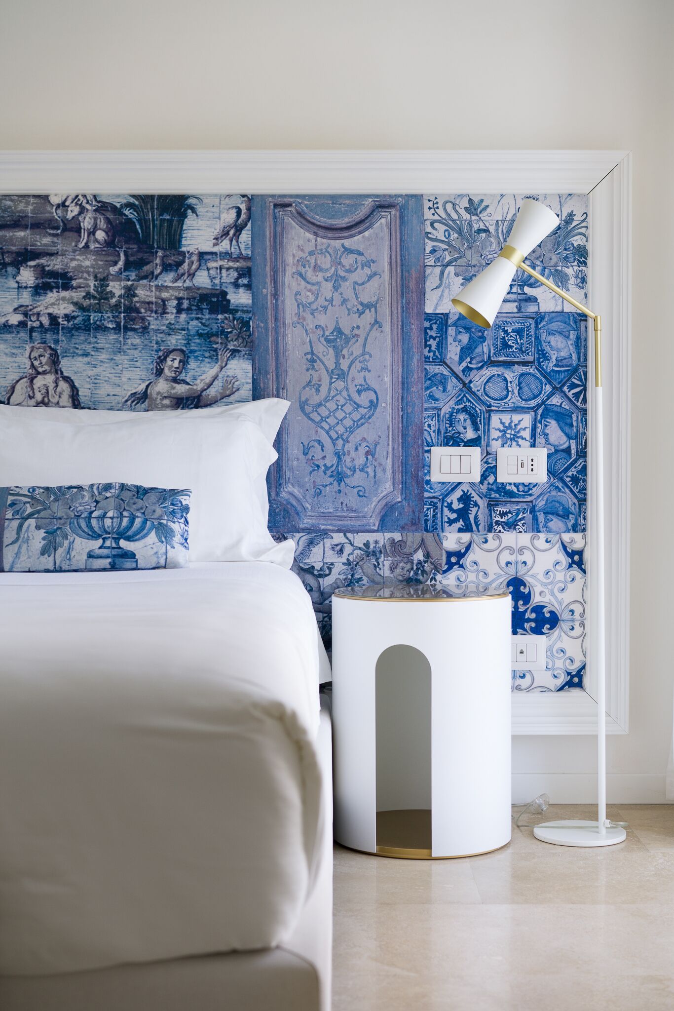 A bedroom with a bed, a white bedside table, a floor lamp, and a wall with blue and white patterned tiles.