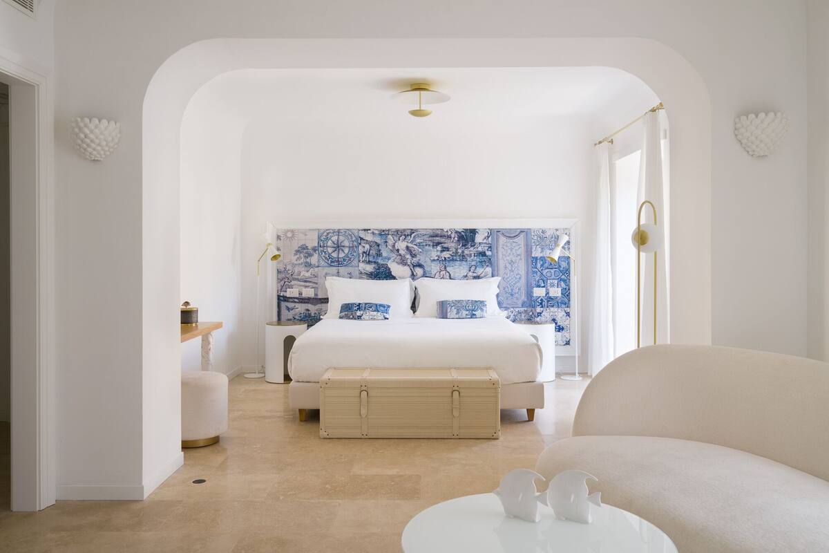 A bedroom with a large bed, a bedside table, a lamp, a suitcase, and a chair.