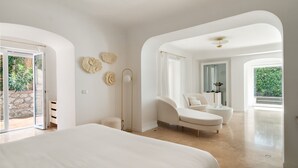 A spacious bedroom with a large bed, a sitting area with a chair and a small table, and a view of the outdoors through a glass door. - La Residenza Capri (Capri)