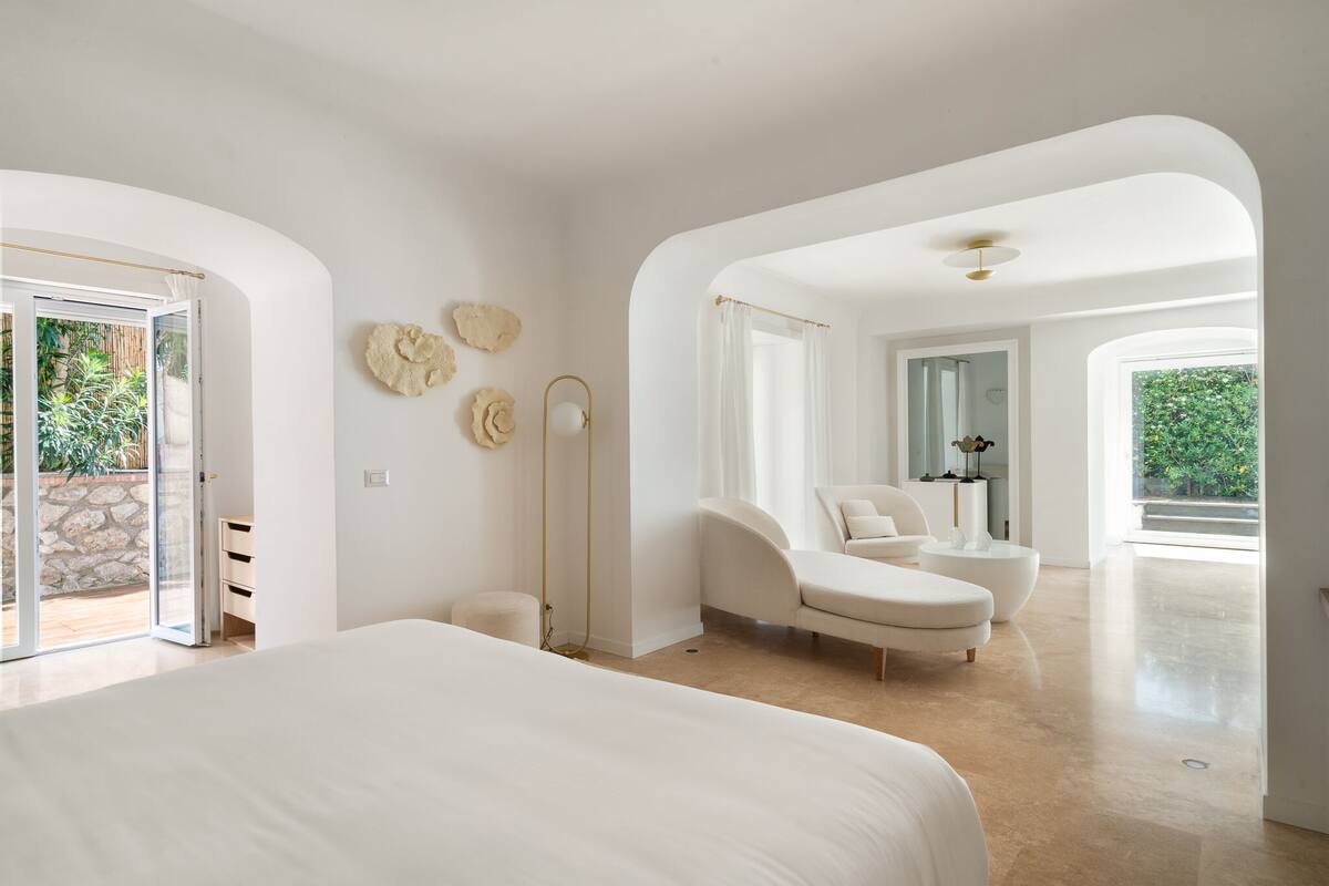 A spacious bedroom with a large bed, a sitting area with a chair and a small table, and a view of the outdoors through a glass door.