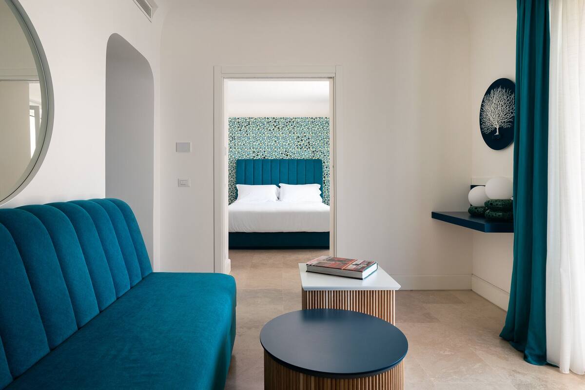 A modern hotel room with a teal sofa, a round wooden side table, a bed with a blue headboard, and a wall-mounted shelf with decorative items.