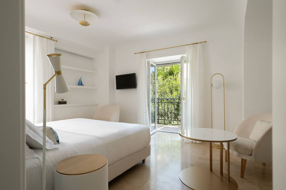 A modern hotel room with a large bed, a round table, a chair, a TV, and a balcony with a view of greenery.