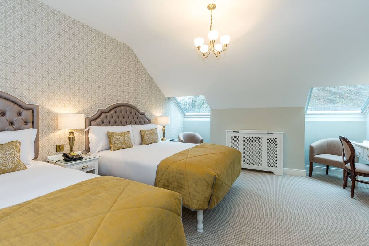A hotel room with two beds, a chandelier, a desk with a lamp, a chair, and a radiator.