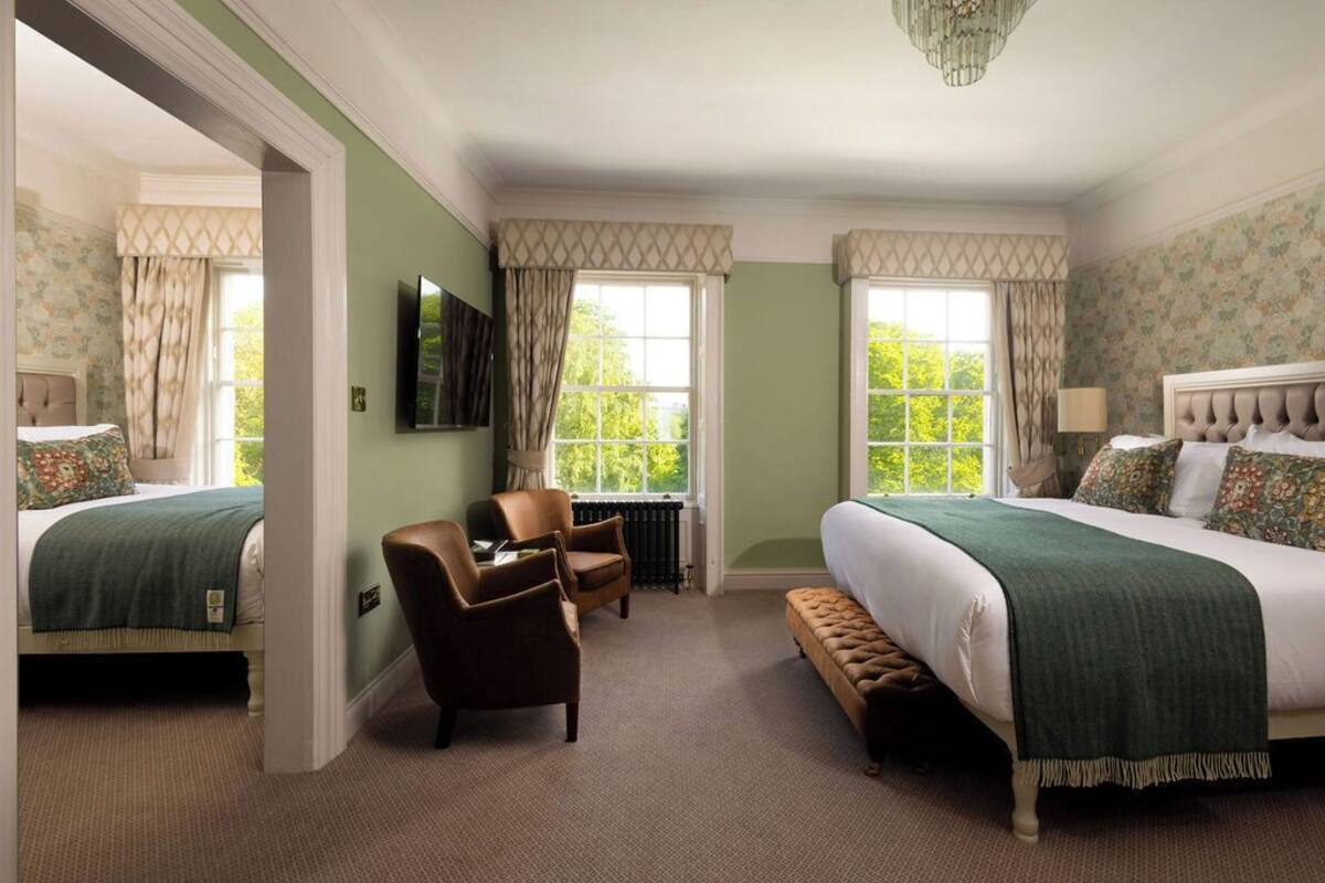 A hotel room with two beds, a sitting area with a chair, a bench, and a large window providing a view of greenery.