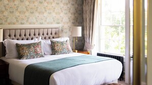 Un dormitorio con cabecera capitonada, papel tapiz floral, una cama grande con muchas almohadas, un banco al pie de la cama, una araña y una ventana con cortinas.