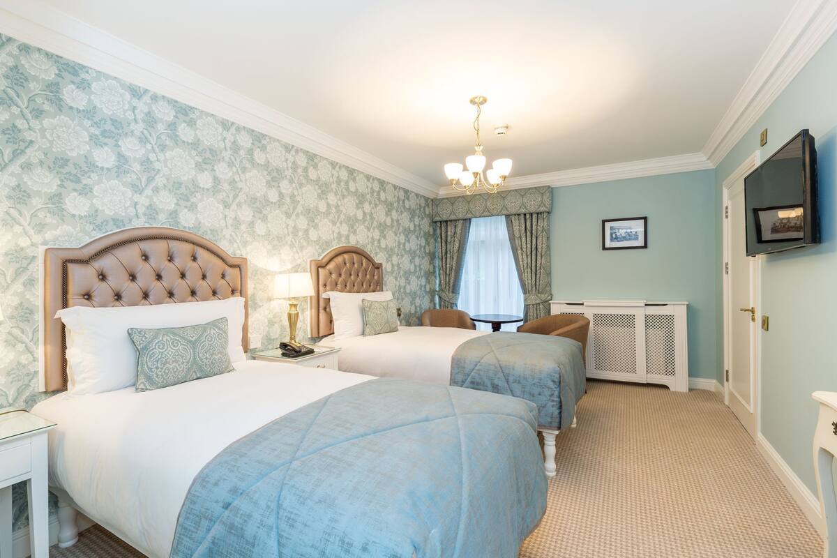 A hotel room with two beds, a television, a chandelier, and a window with curtains.