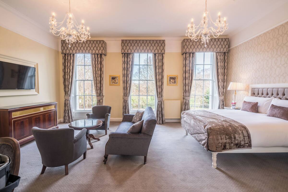 A hotel room with a large bed, a sofa, a chair, a TV, and a chandelier.