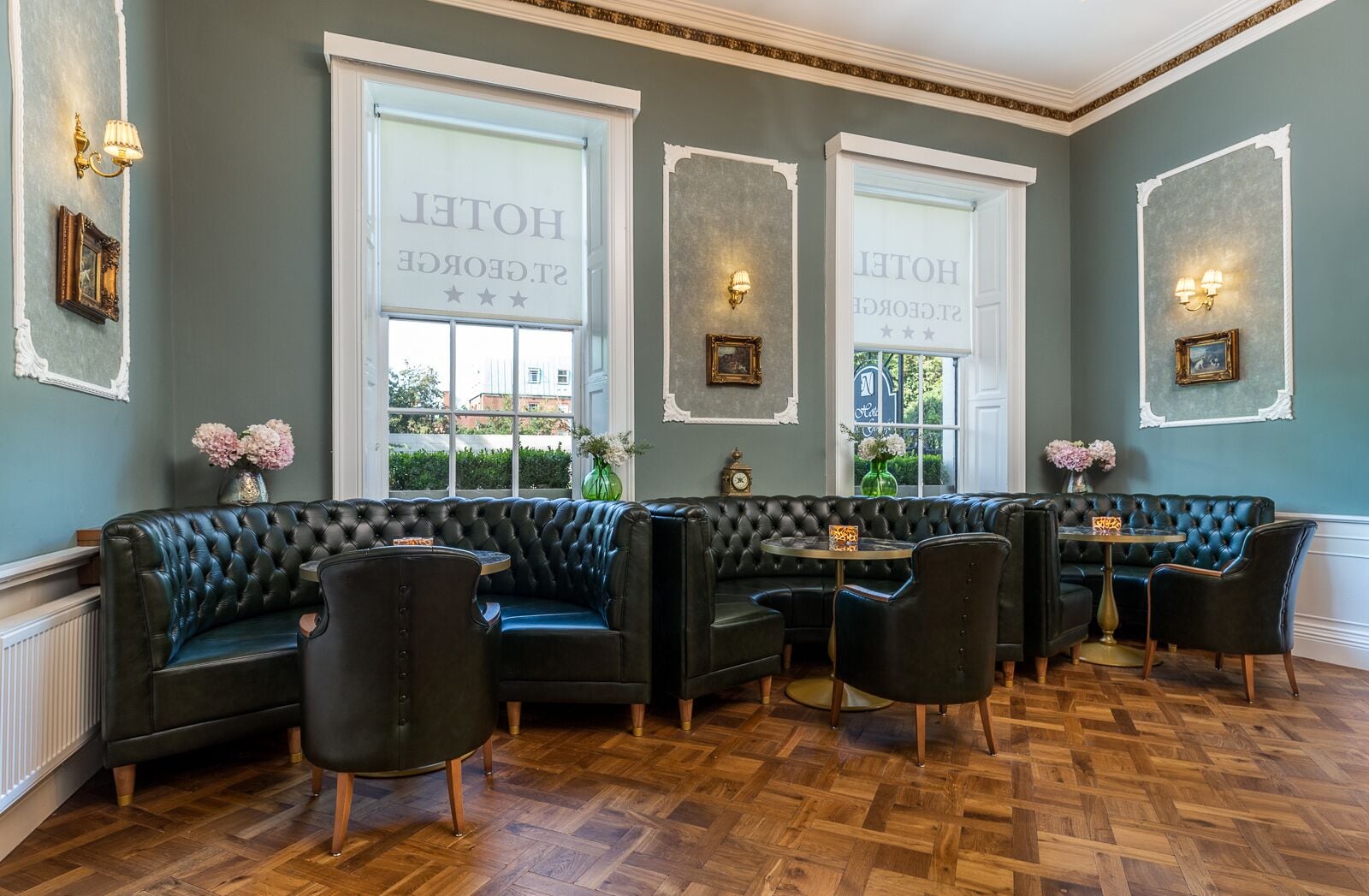 En hotellobby med klassisk design, med en sort lædersofa og lænestole, parketgulv i træ og store vinduer med hotellets navn.