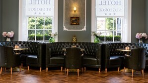 Eine Hotellobby mit einem Kronleuchter, zwei großen Fenstern mit der Beschriftung 'HOTEL ST. GEORGE', einem schwarzen Ledersofa und einem grünen Strauß in einer Vase.