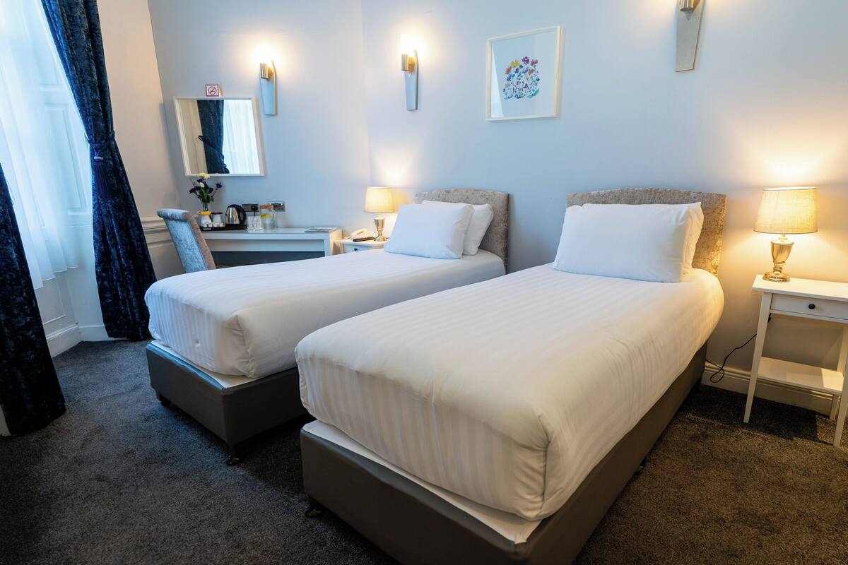 A hotel room with two beds, a desk, a chair, a mirror, and a small table with a lamp.