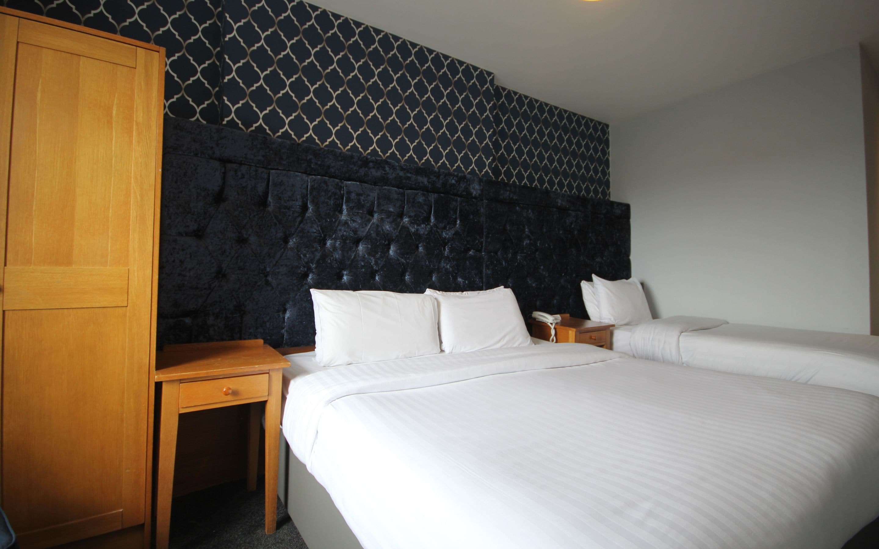A hotel room with two beds, a wooden wardrobe, a small bedside table, and a patterned wall.