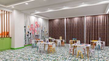 Children can play and learn in this colorful playroom with a patterned carpet, tables, and chairs, surrounded by a vibrant mural.