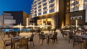 An outdoor dining area with tables and chairs, a pool, and a building with a unique geometric facade. - Wyndham Bukhara (Bukhara)