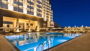 A swimming pool with lounge chairs and umbrellas, surrounded by a building with a modern design and multiple windows. - Wyndham Bukhara (Bukhara)