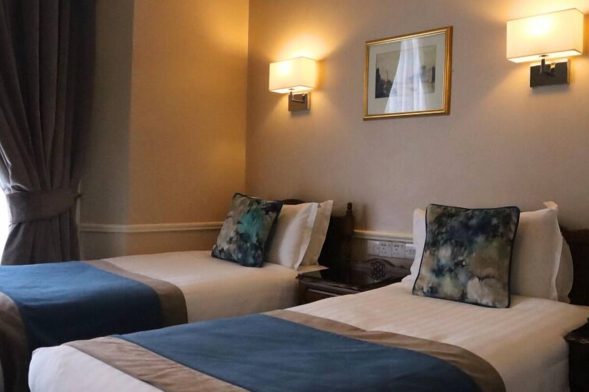 Une chambre d’hôtel avec deux lits, chacun recouvert d’une couverture bleue et agrémenté d’un oreiller orné d’un motif abstrait bleu et vert. Deux lampes murales sont fixées de part et d’autre d’un tableau encadré.