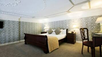 A bedroom with a four-poster bed, two bedside tables with lamps, a wooden chair, and patterned wallpaper.