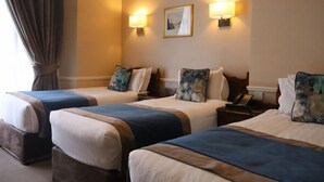 A hotel room with three beds, each with a blue and beige bedspread, and matching pillows. There are two wall-mounted lamps and a framed picture above the beds.