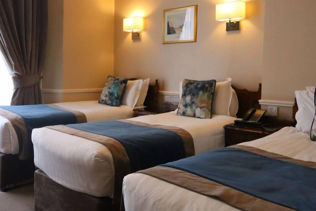 Une chambre d’hôtel avec trois lits, chacun recouvert d’une courtepointe bleue et beige, et agrémentée d’oreillers assortis. Deux lampes sont fixées au mur et un tableau encadré est accroché au-dessus des lits.