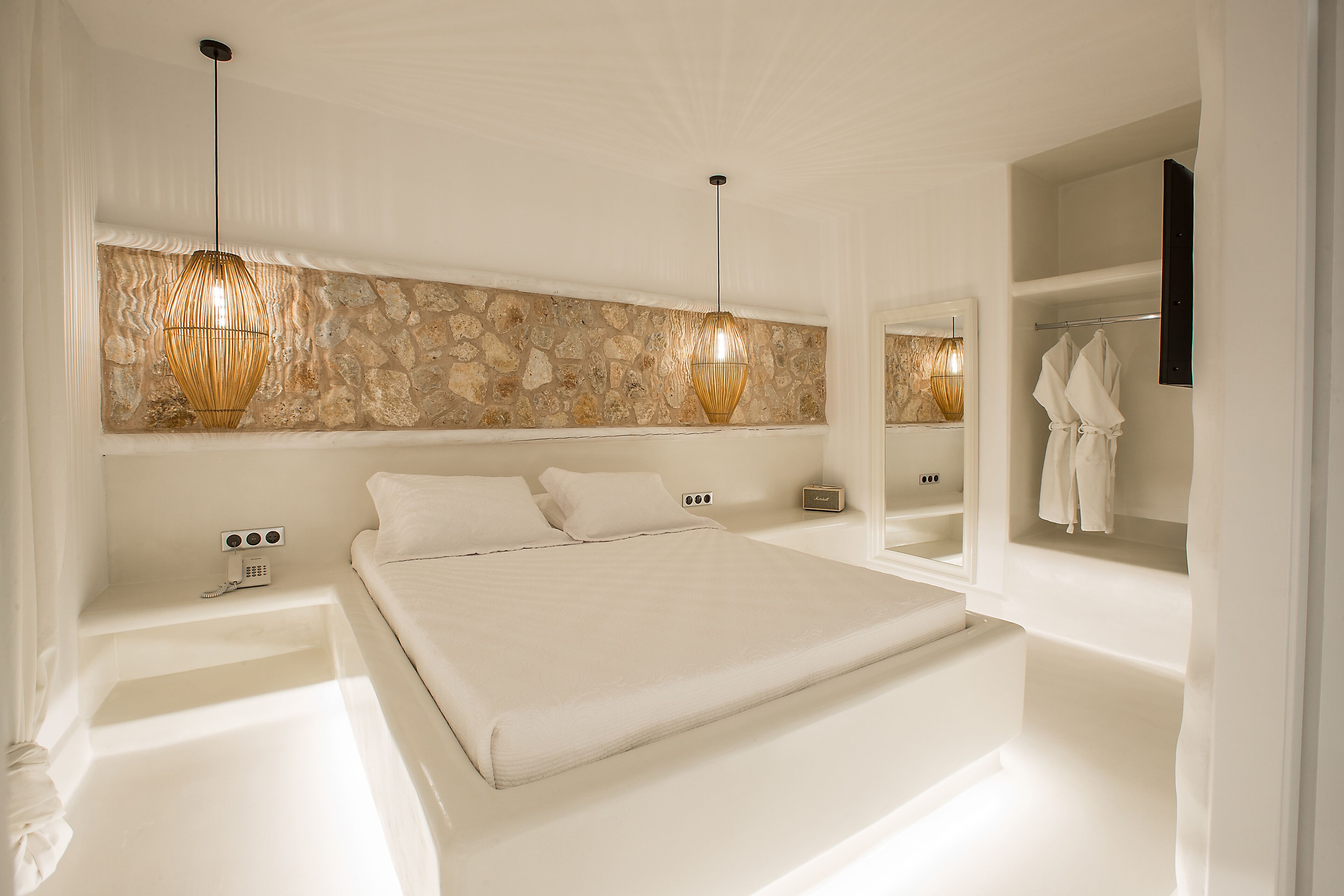 Une chambre moderne avec un grand lit, un mur d’accent en pierre et une coiffeuse intégrée dotée d’un miroir et de crochets pour les peignoirs.