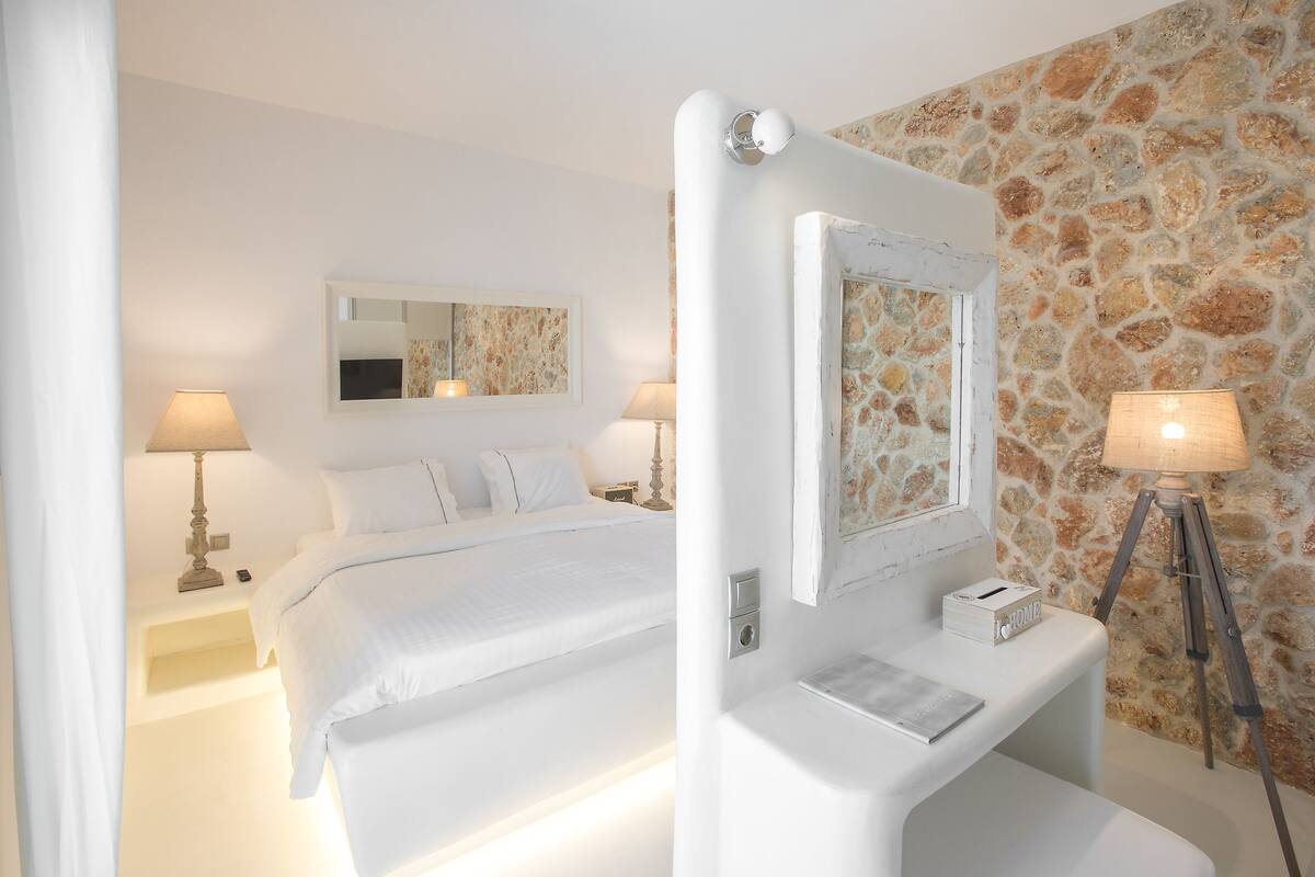 A hotel room with a bed, bedside lamps, a mirror, a bedside table with a lamp, and a stone wall.