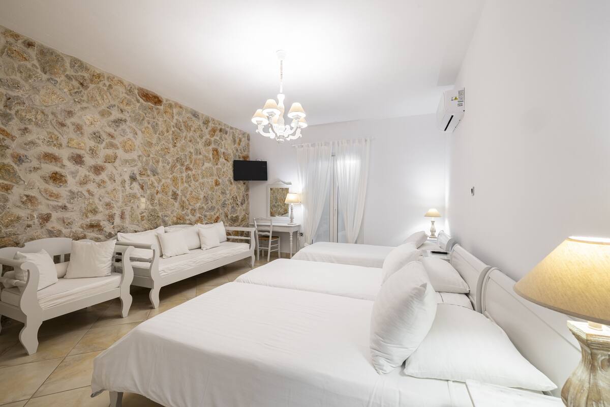 A hotel room with a stone wall, white furniture, a chandelier, a flat-screen TV, and a bed with white bedding.