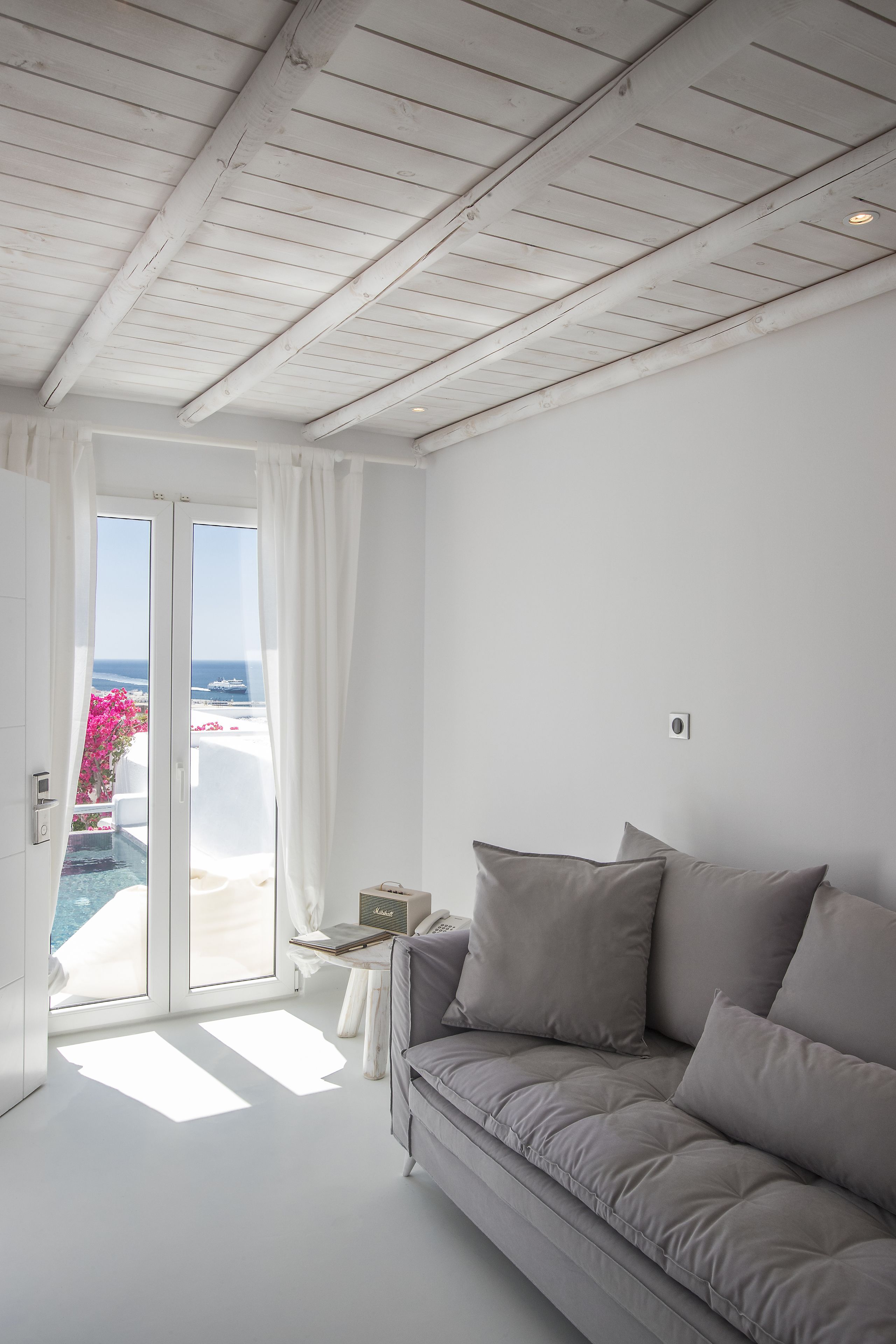 Ein modernes Zimmer mit weißer Decke, einem grauen Sofa und einer Schiebeglastür, die auf einen Balkon mit Meerblick führt.