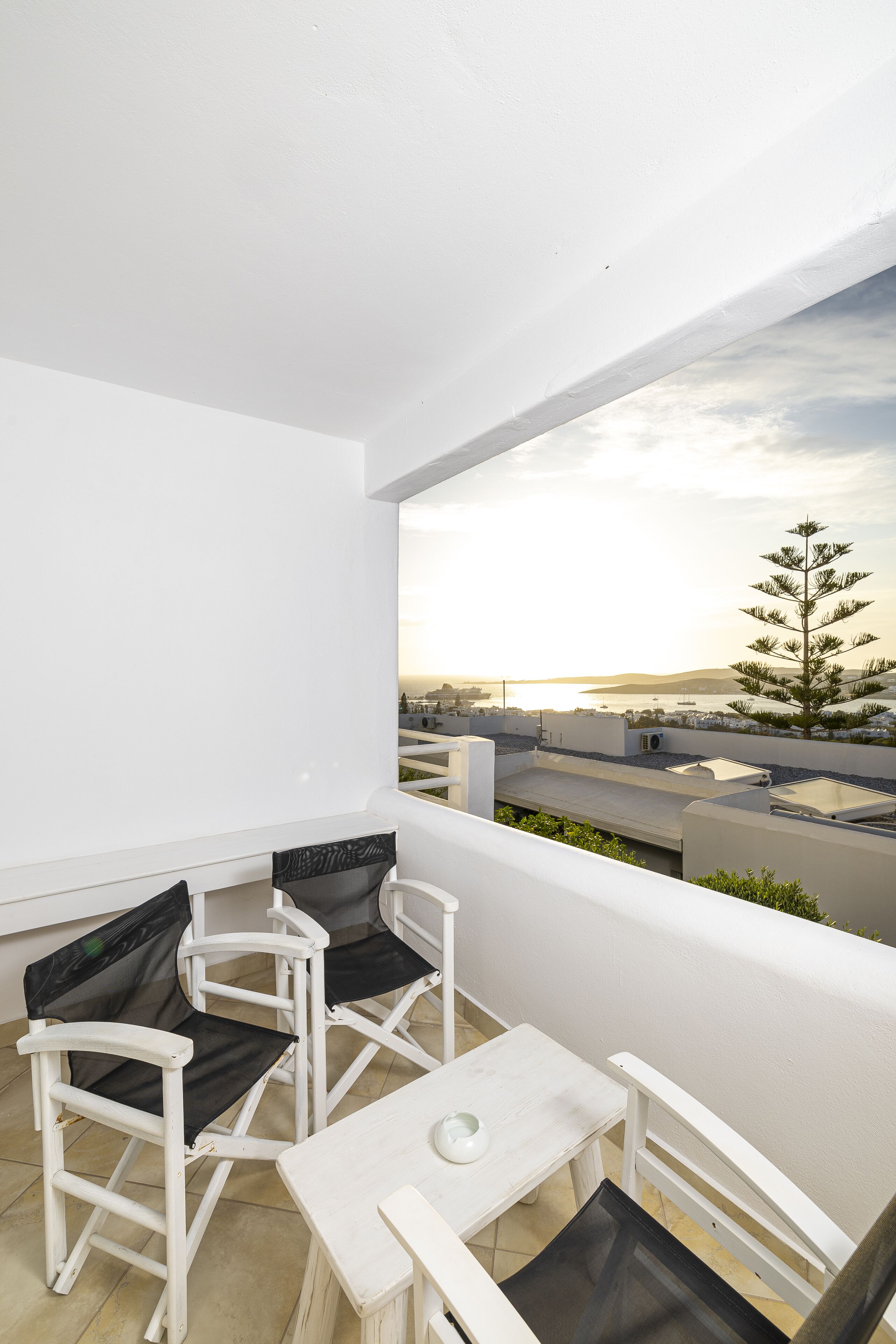 A balcony with white walls, two white chairs with black cushions, a small white table, and a view of a coastal landscape with a tree and buildings.