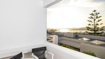 A balcony with white walls, two white chairs with black cushions, a small white table, and a view of a coastal landscape with a tree and buildings.