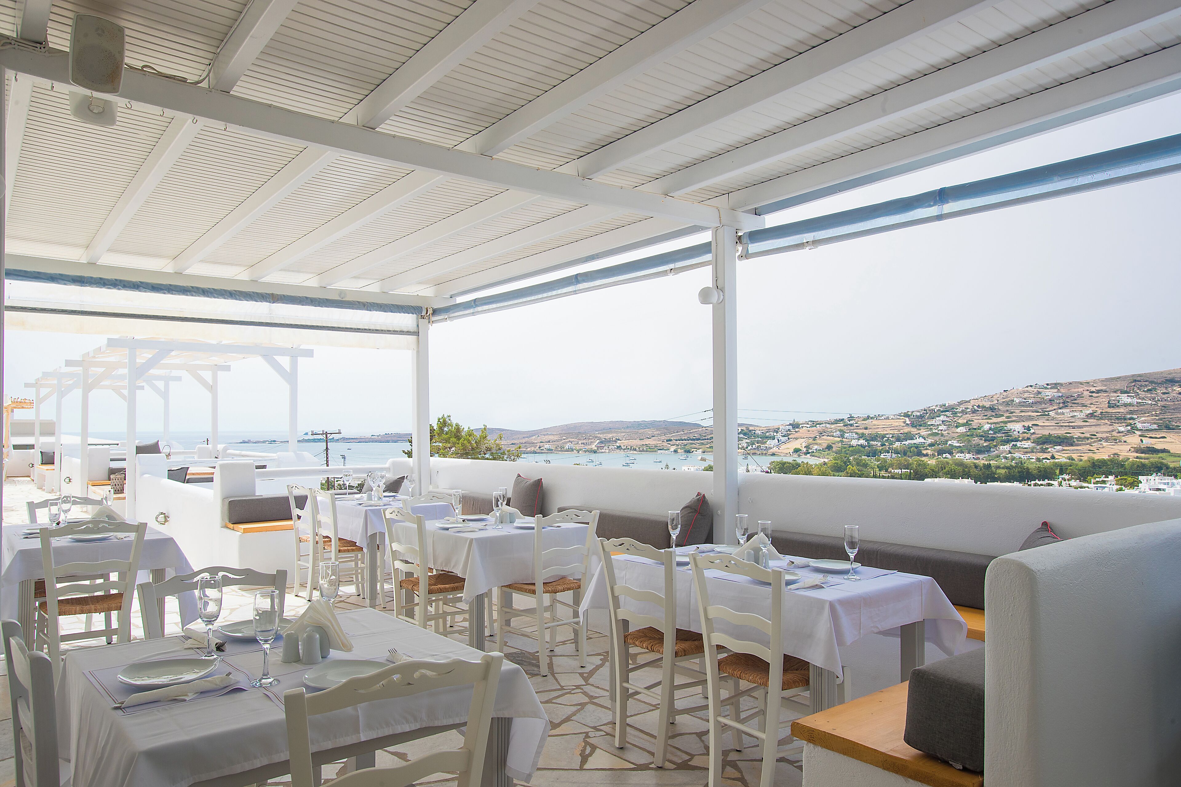 An outdoor dining area with white tables and chairs, overlooking a scenic view of the sea and hills.