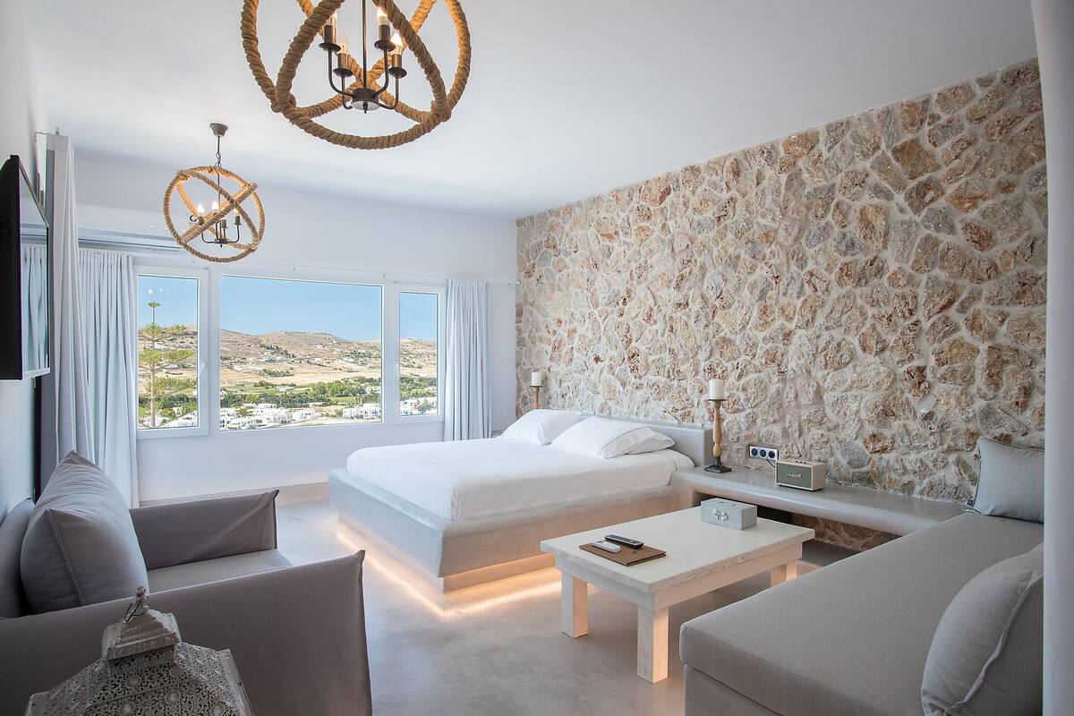 A modern bedroom with a large bed, a stone accent wall, a view of the landscape, and a chandelier.