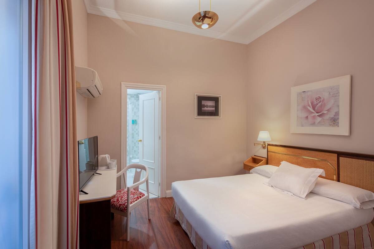 A hotel room with a bed, a desk, a chair, a desk lamp, and a framed picture on the wall.