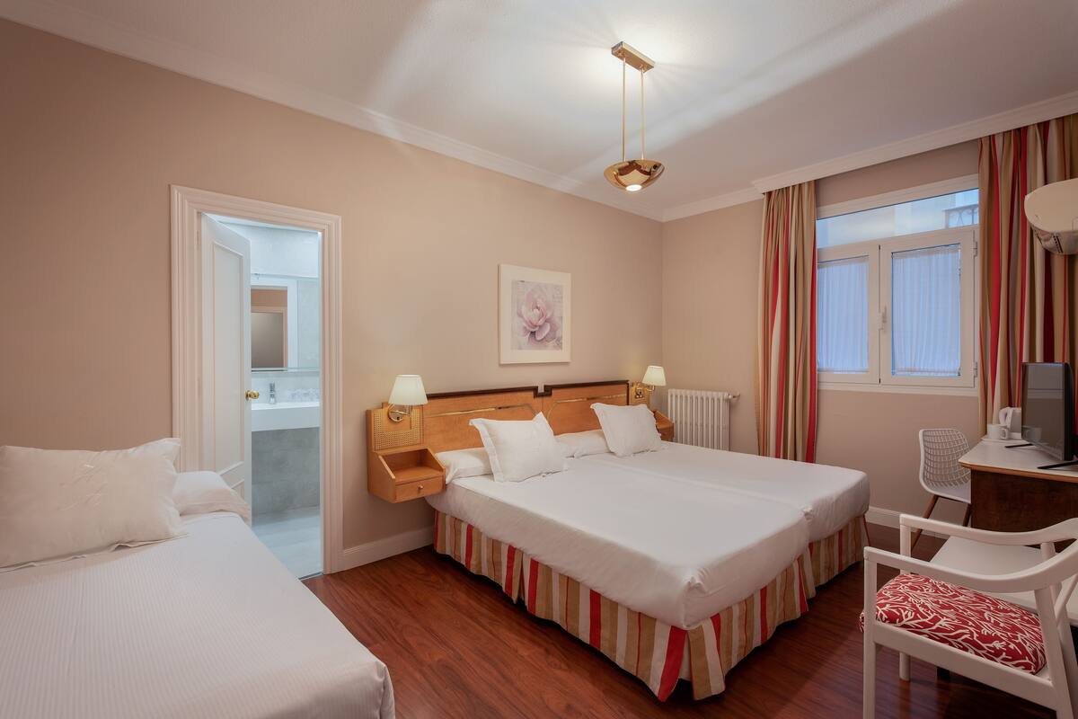 A hotel room with two beds, a wooden headboard, a desk with a chair, a radiator, and a window with curtains.